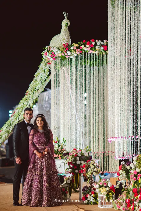 Bride wearing wine gown and groom wearing black tuxedo for the reception posing against a gigantic ombre wall of pink blooms